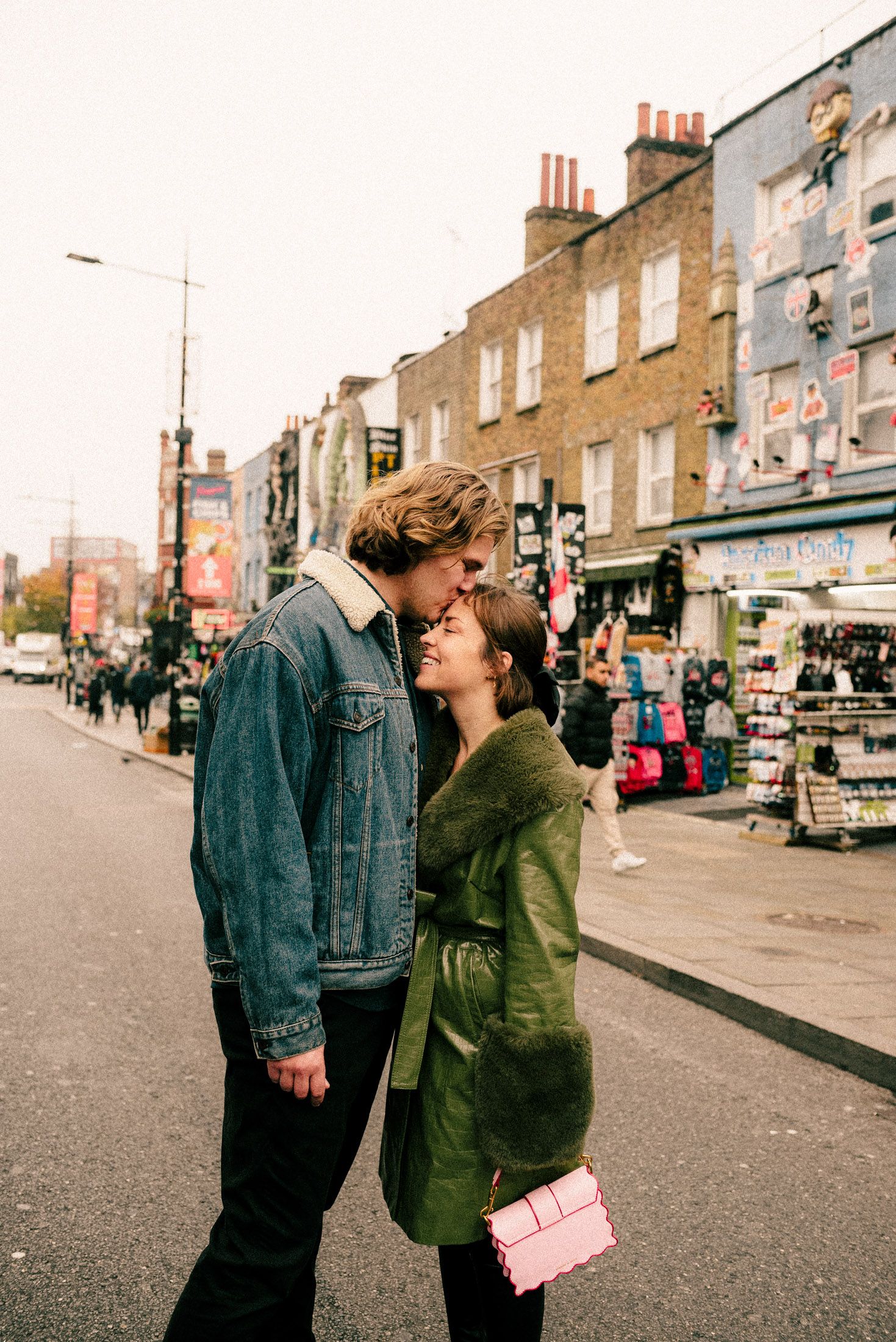 A couple shares an intimate moment on a city street with brick buildings and storefronts in the background.