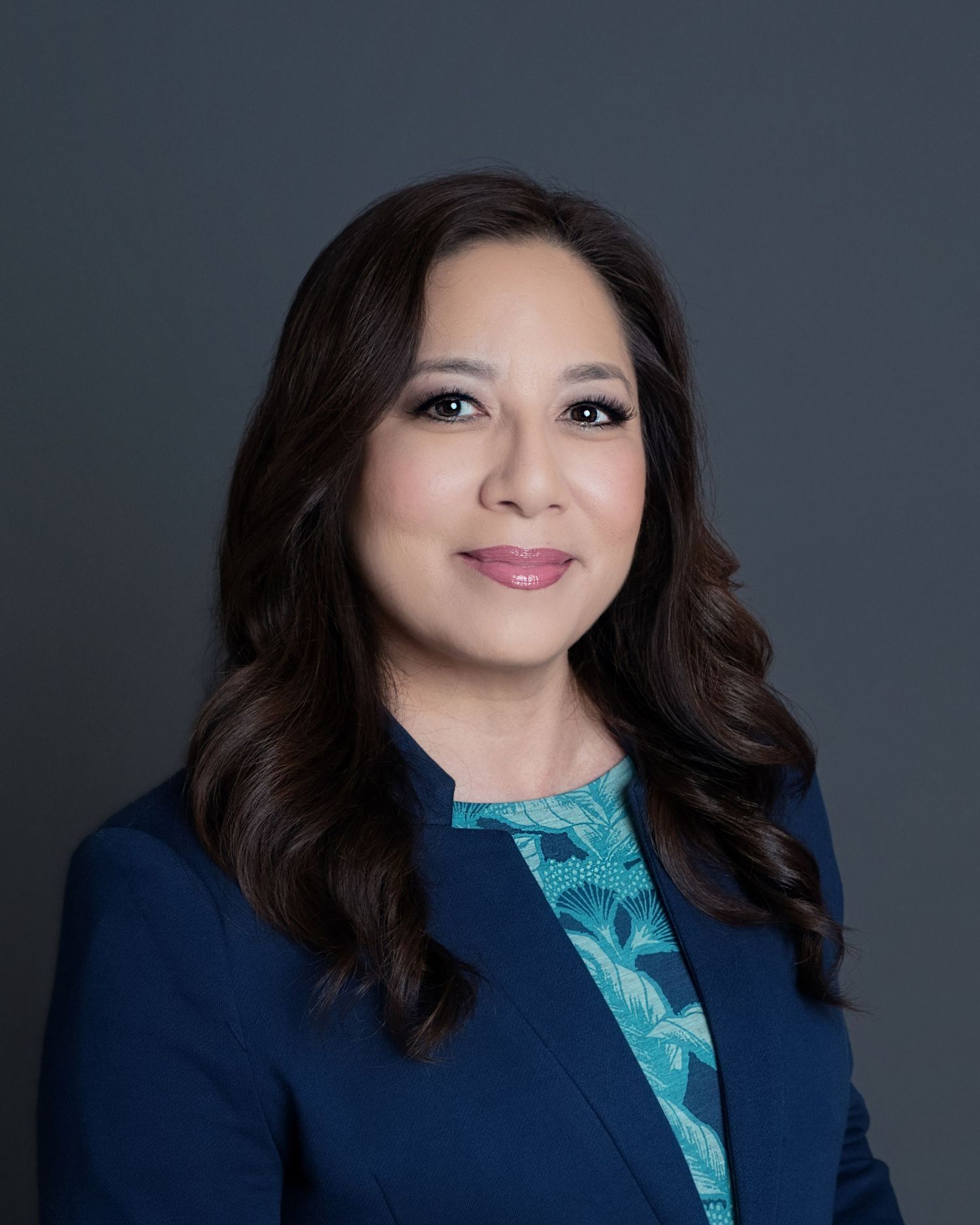 Professional headshot of a person wearing a navy blazer and teal patterned blouse against a dark gray background.