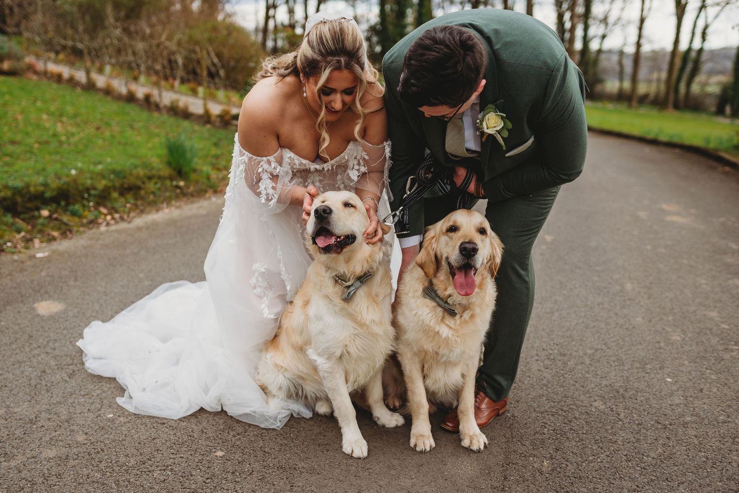 A couple in wedding attire poses with their two golden retriever dogs in an autumn park setting.