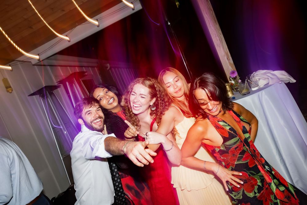 Wedding guests dance and celebrate on a colorful dance floor illuminated by purple and red lighting at a reception party.
