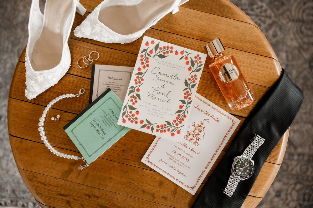Wedding details including invitations with floral design, white heels, perfume bottle, and jewelry arranged on wooden table.