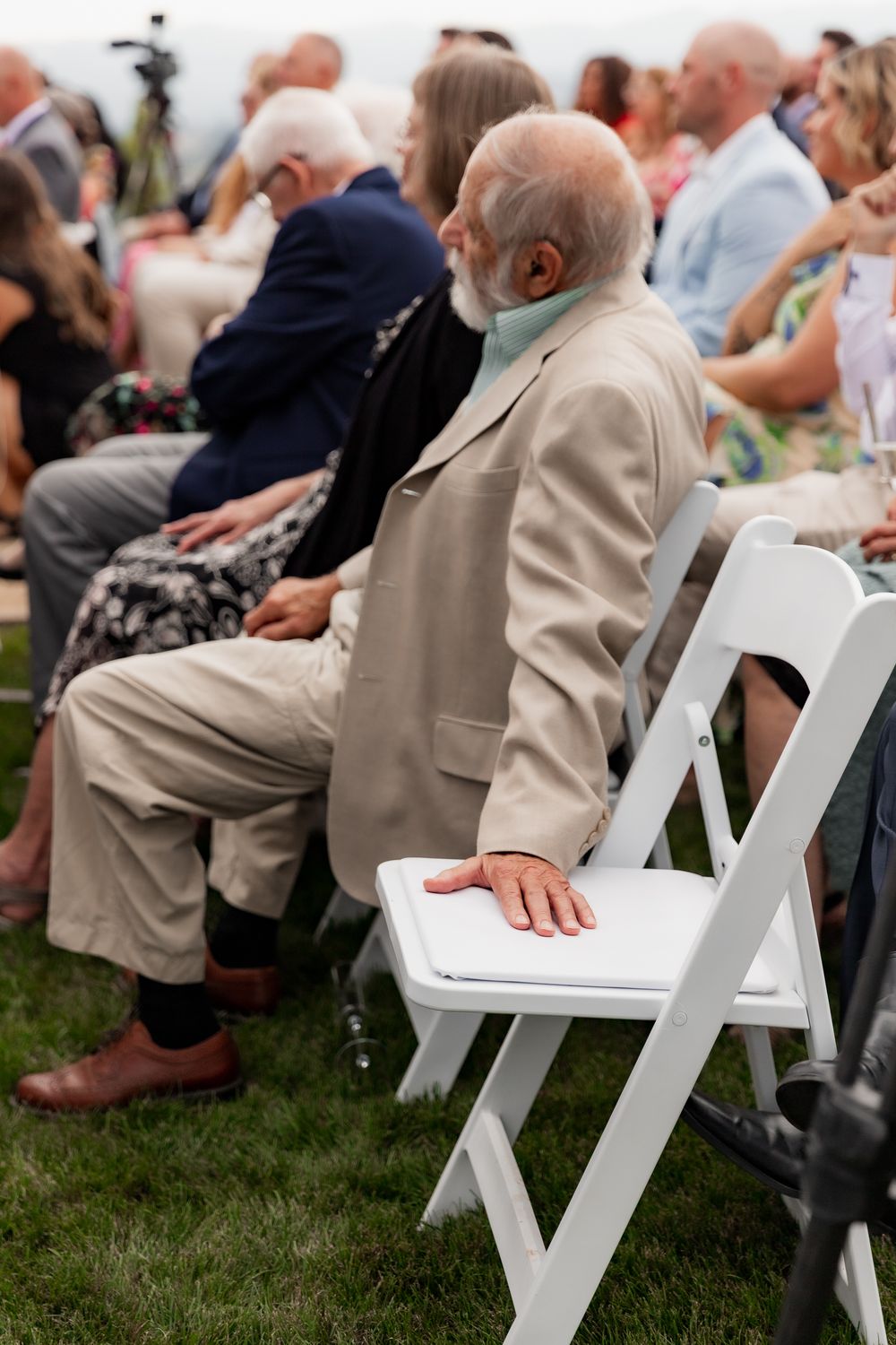 People seated in white folding chairs on grass at an outdoor event, with focus on hands resting on chair armrests.