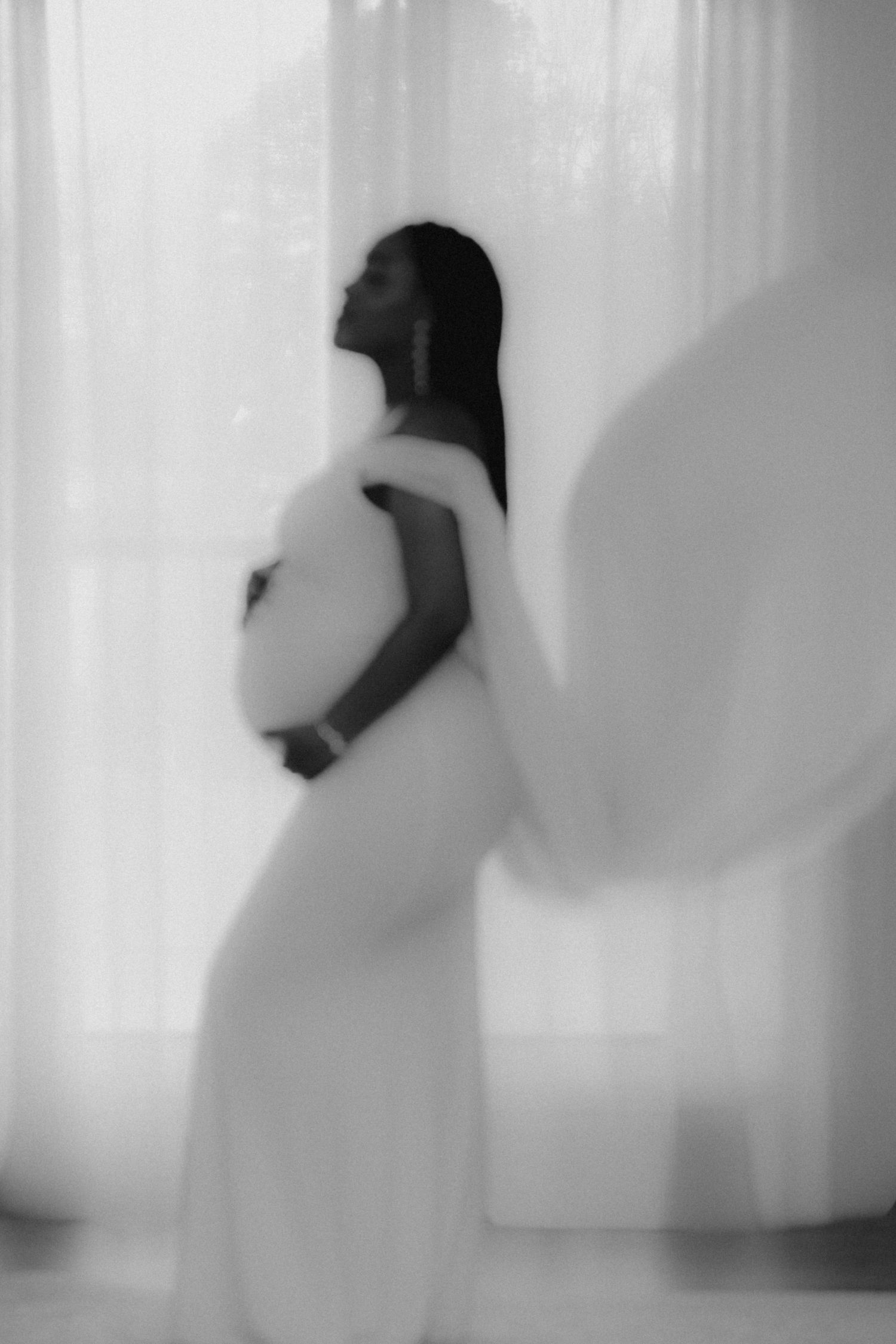 Artistic black and white silhouette of a pregnant woman in a flowing dress standing against sheer curtains.