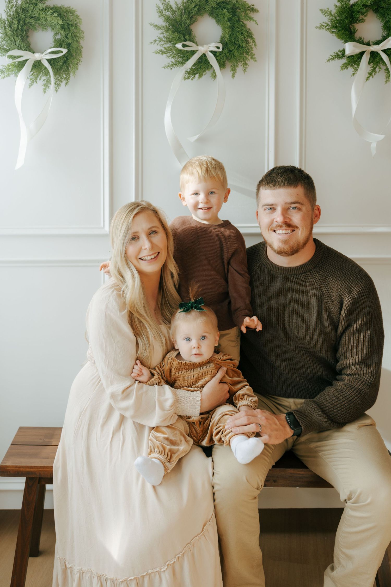 A family wearing coordinated cream and brown outfits poses together against a white wall decorated with evergreen wreaths.