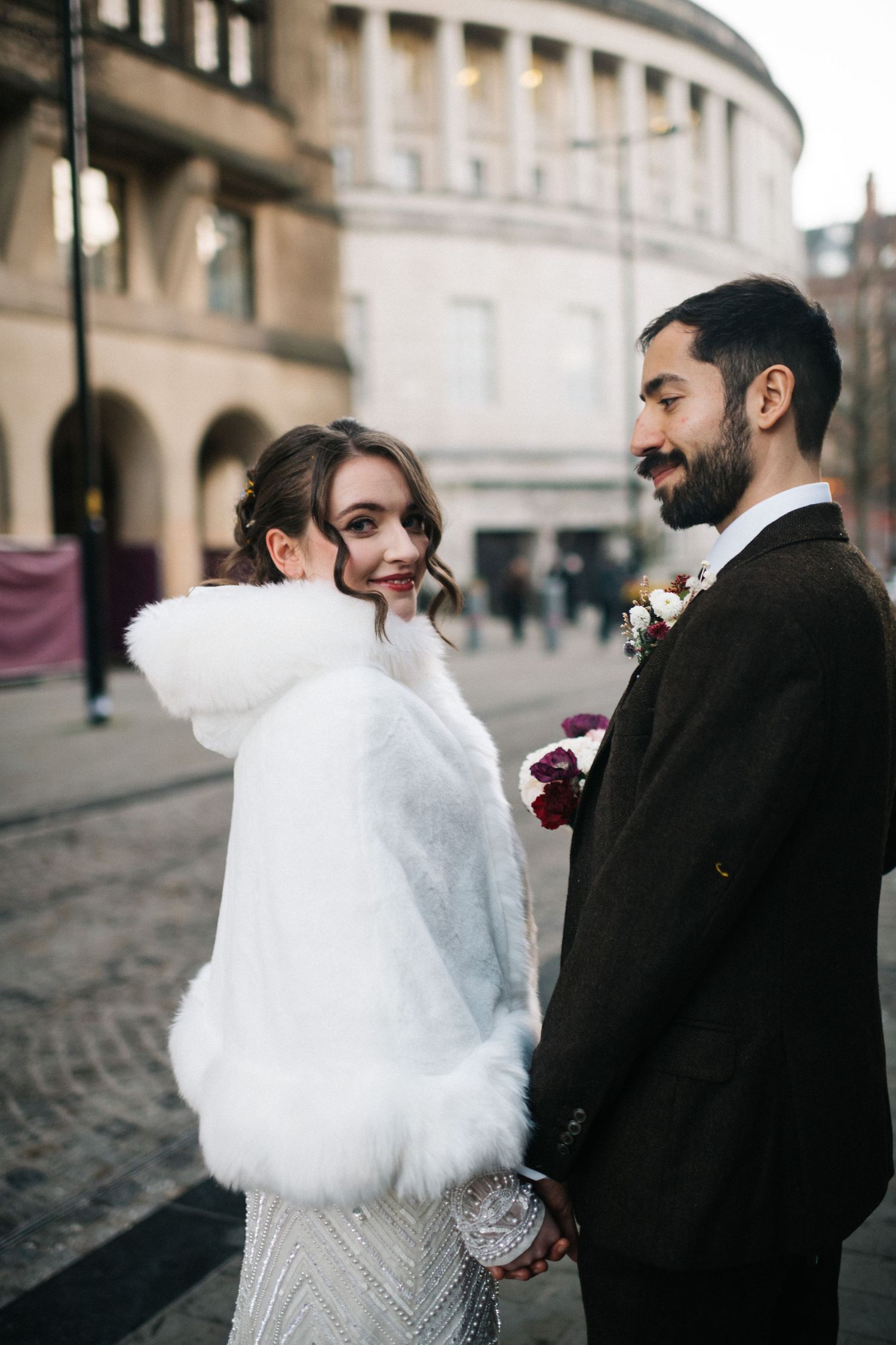 Romantic winter wedding couple walking on cobblestone street with bride wearing white fur shawl over gown. Manchester City centre wedding.