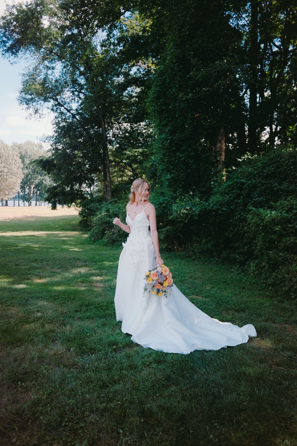 A bride in a flowing white gown poses gracefully in a natural outdoor setting with trees and sunlit grass.