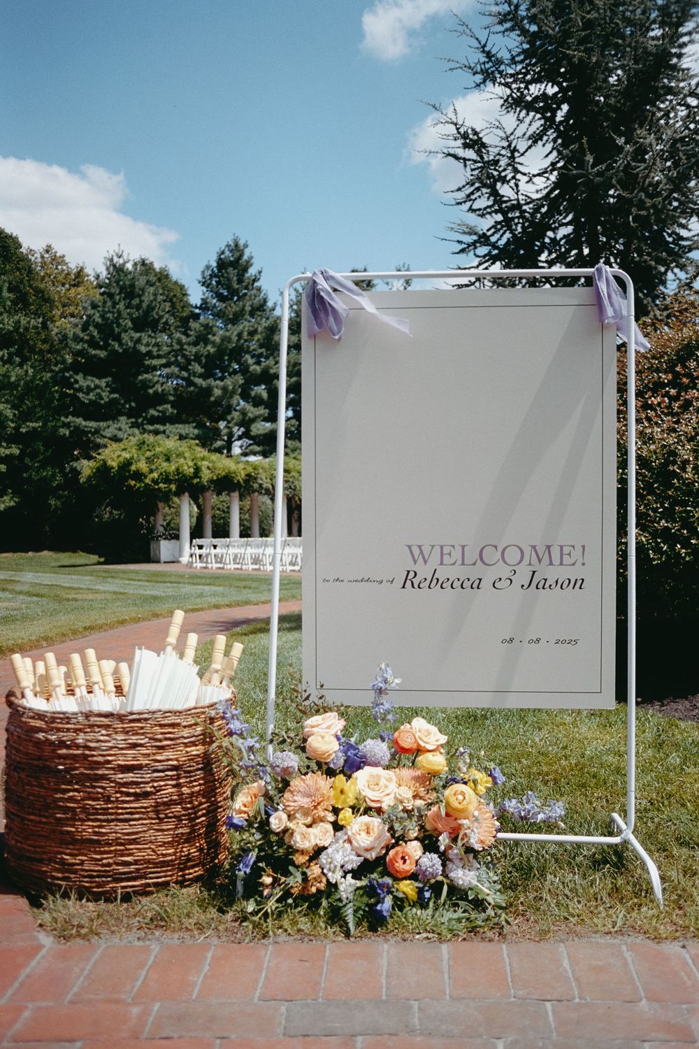 Outdoor wedding welcome sign decorated with rustic woven basket and colorful floral arrangements on brick patio.