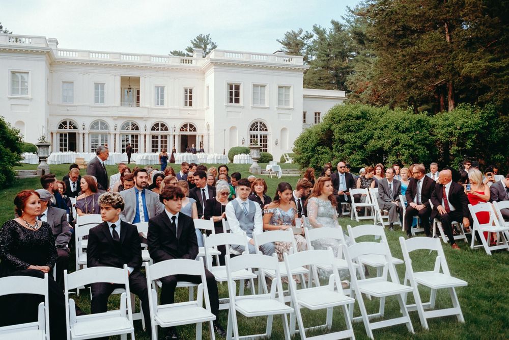 Elegant outdoor wedding ceremony setup with white chairs on a lawn in front of a grand white mansion.