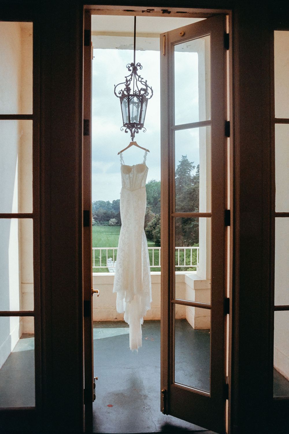 White wedding dress hangs elegantly from ornate chandelier in doorway framed by French doors overlooking balcony.