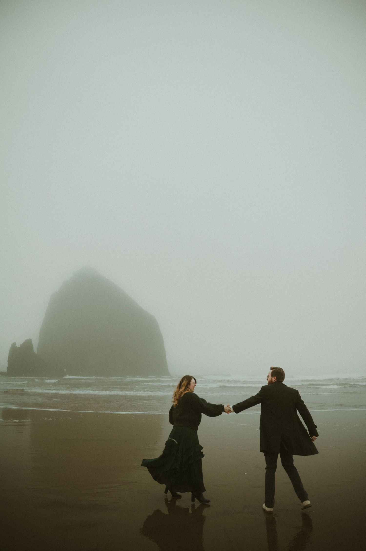 Two silhouettes walk along a misty beach with dramatic rock formations emerging from the fog in the background.