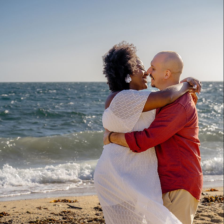 Romantic couple embraces on sunny beach with ocean waves and sparkling water in the background.