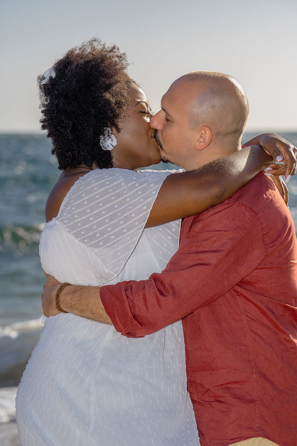 A couple embraces and kisses on the beach at sunset, with the ocean waves visible in the background.