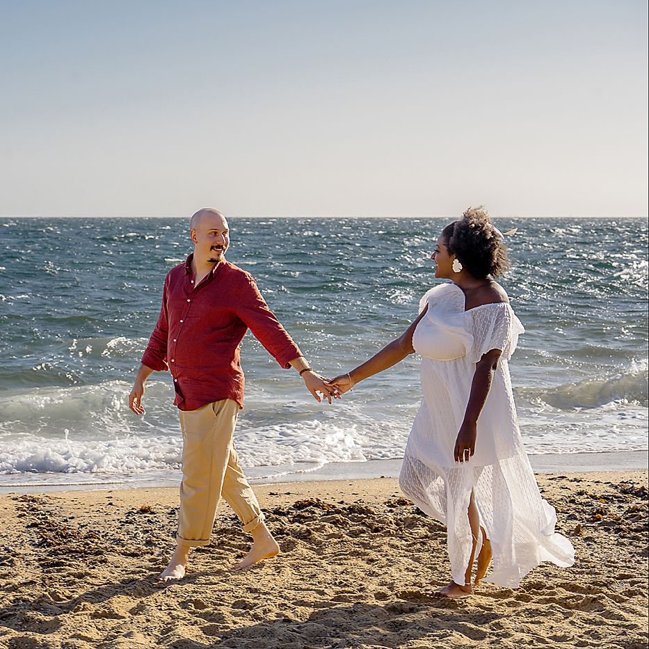 Two people walking hand in hand along a sunny beach with ocean waves crashing in the background.