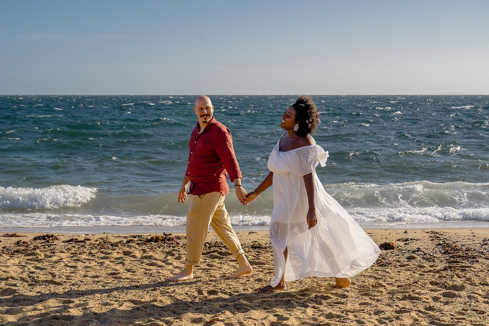 A couple walks hand in hand along a sunny beach with waves crashing behind them.
