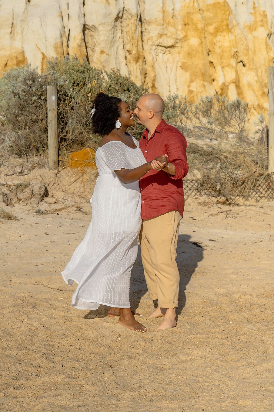 A couple dances together on sandy ground against a weathered yellow wall backdrop during a sunny day.
