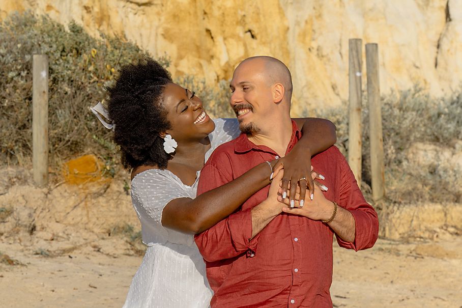 A couple laughing and embracing in coordinated outfits against a rustic yellow wall background.