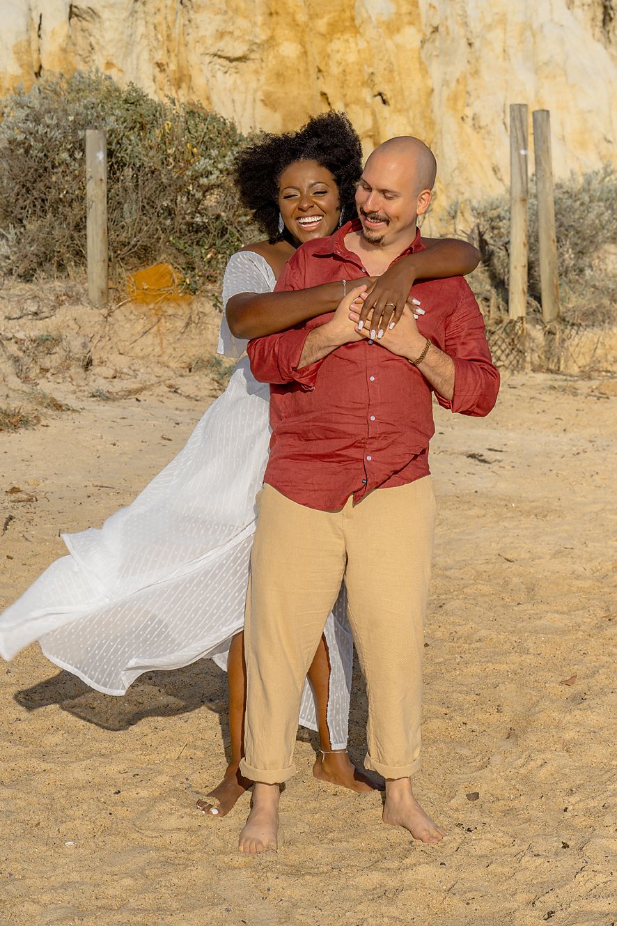 A couple in red and khaki outfits dance together in a romantic outdoor photoshoot against a rustic stone wall.