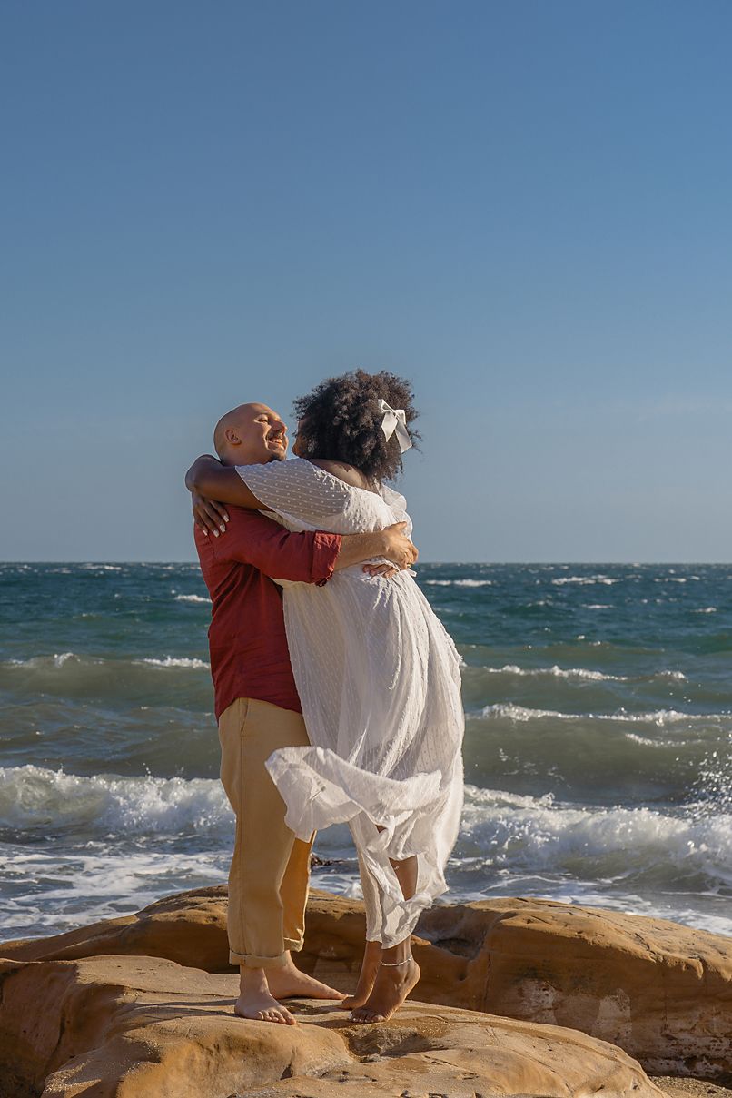 A couple embraces on rocky coastal outcrop with white flowing dress and waves crashing in background on sunny day.