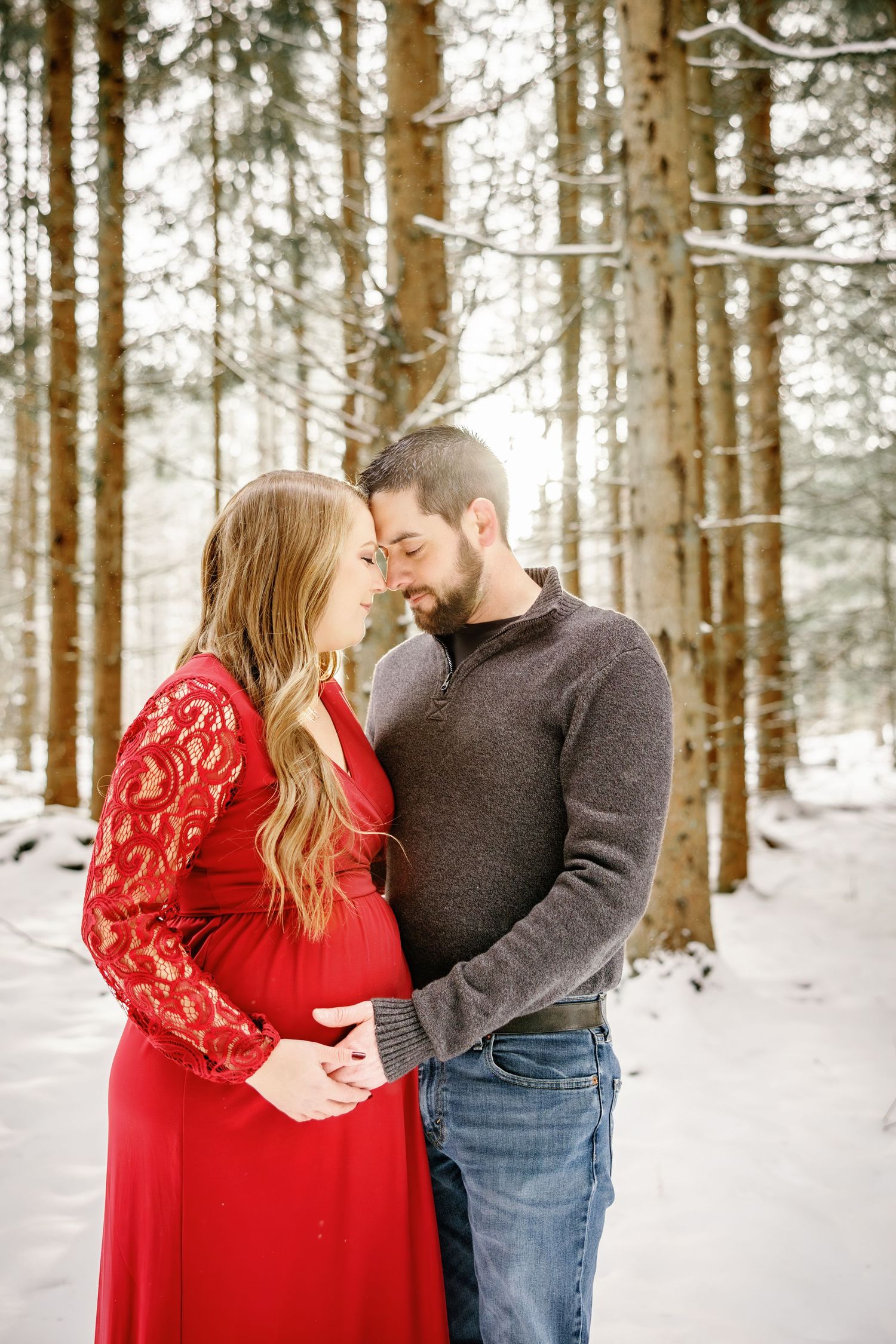 Winter forest maternity photoshoot with couple in red dress and grey sweater among snow-covered pine trees.