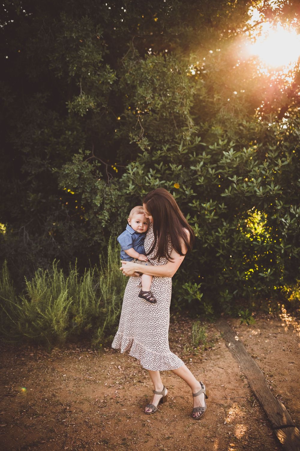 A couple embraces on a dirt path during golden hour with sunlight filtering through trees in the background.