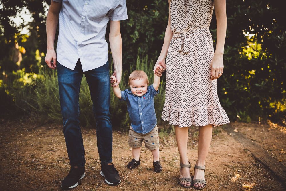 Family wearing coordinated blue and beige outfits stands together on a wooded path during golden hour.