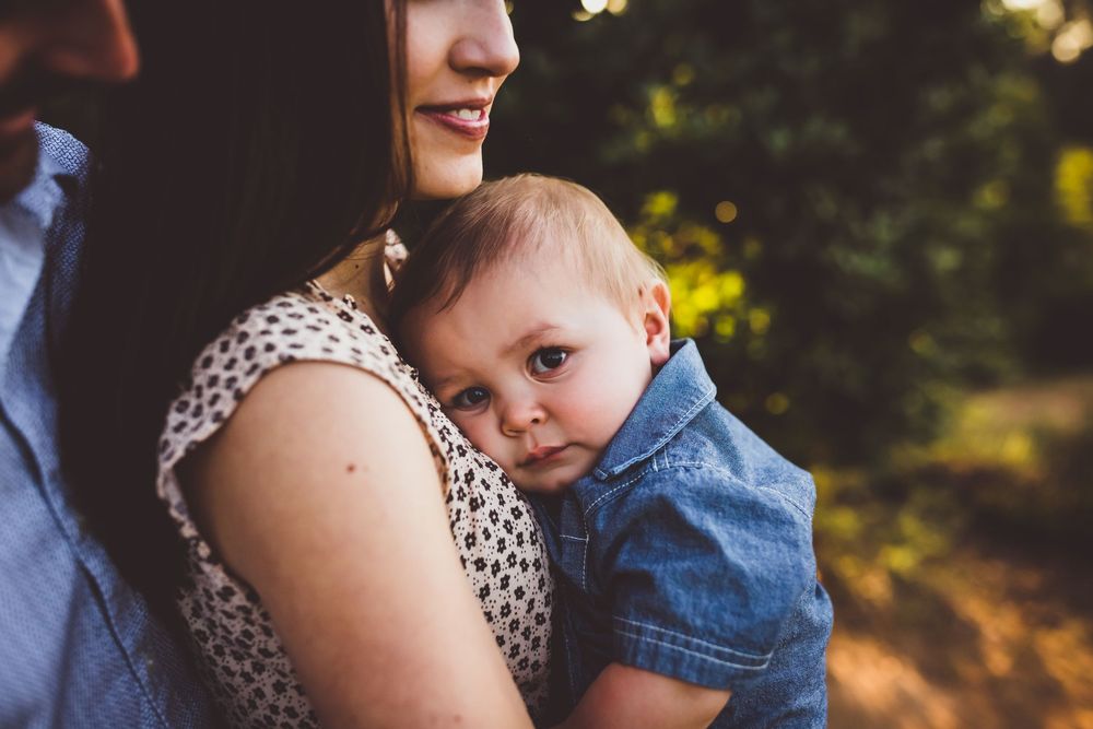 A baby in denim clothing rests against someone wearing a polka dot top in a sunlit outdoor setting.