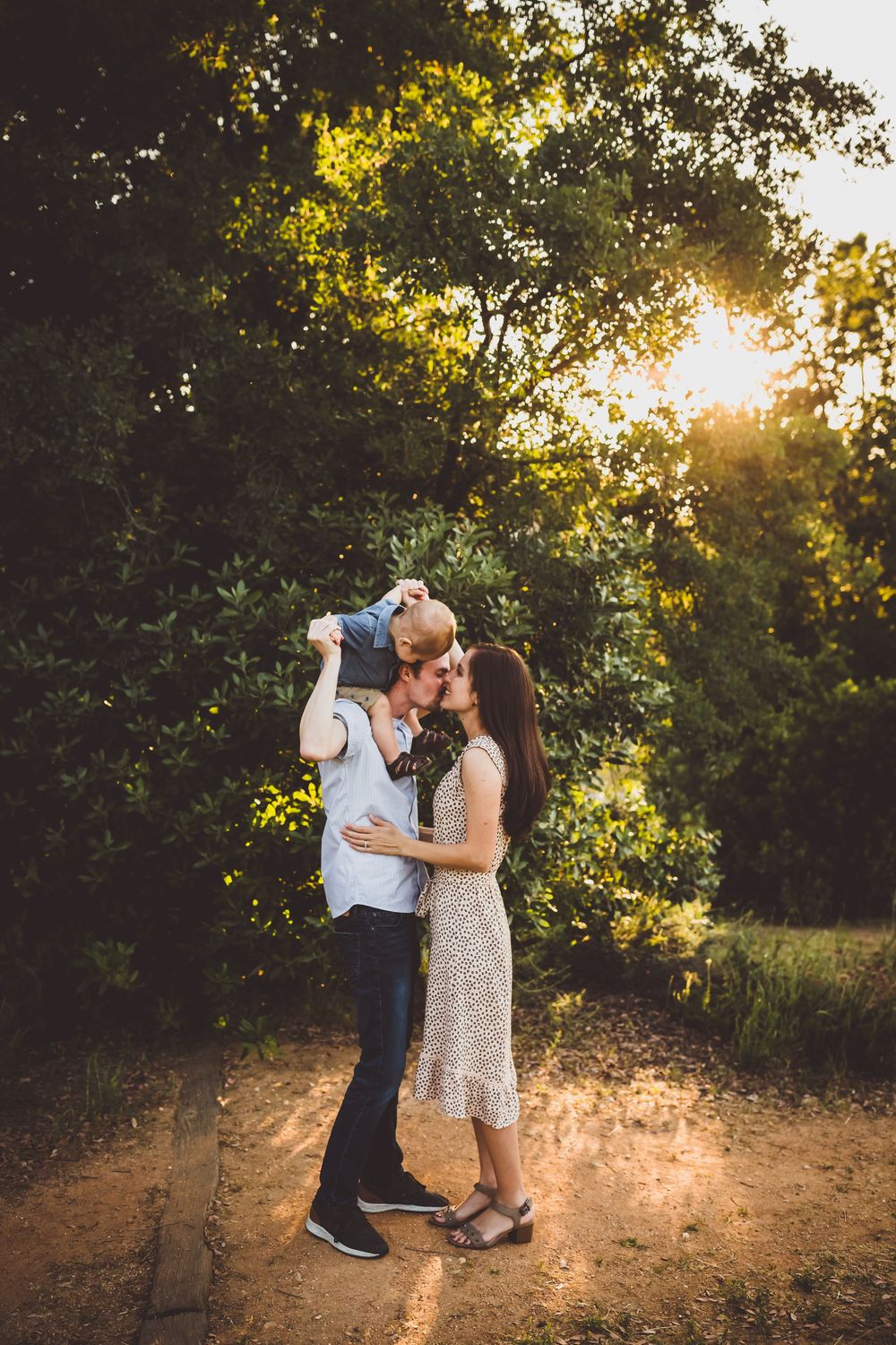 A couple embraces on a sunlit dirt path surrounded by lush trees during golden hour.