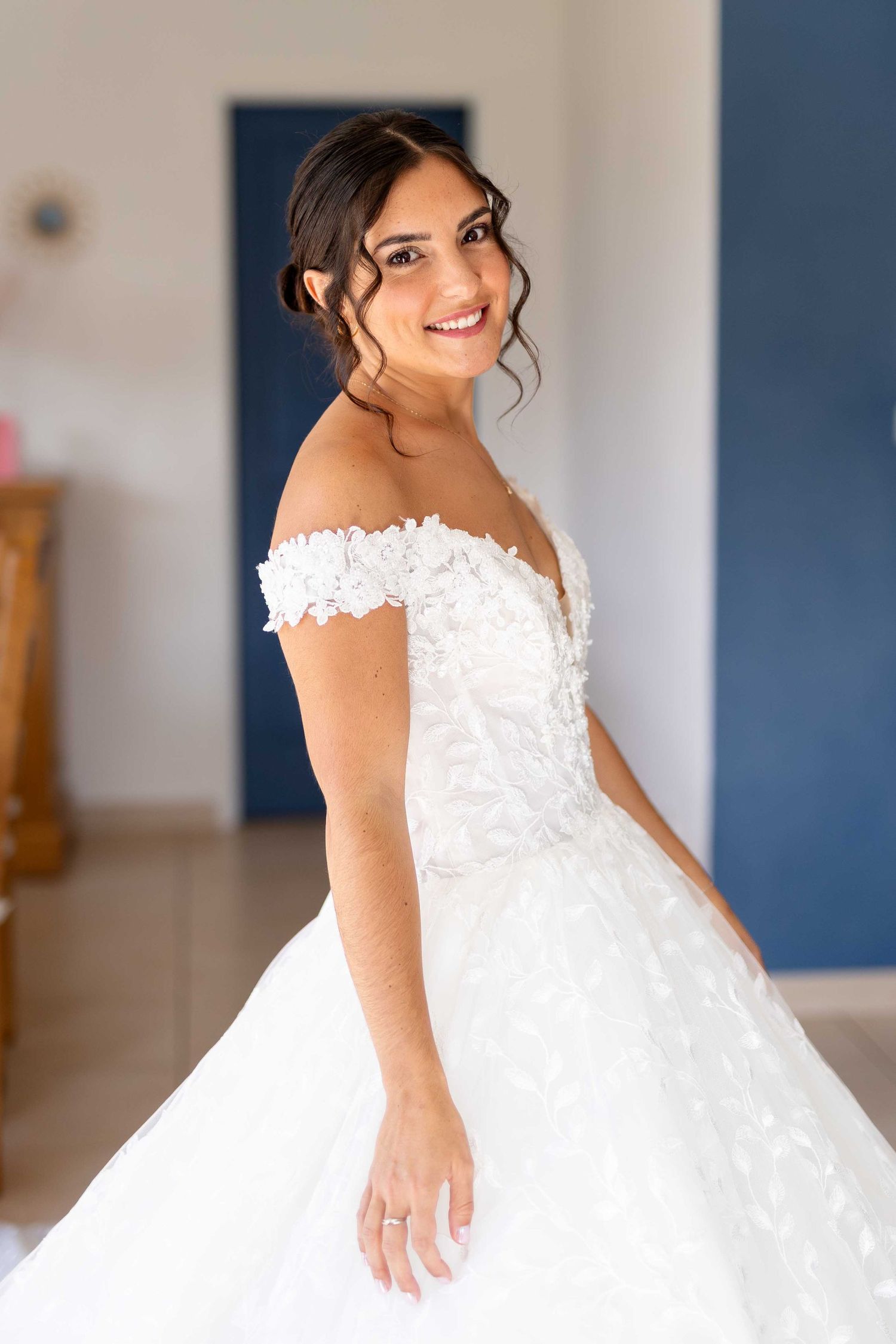 Belle mariée en robe de mariée en dentelle à épaules dénudées avec des cheveux bouclés souriante debout à l'intérieur.