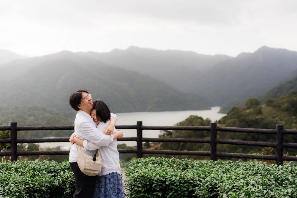 Two people embrace while enjoying a scenic mountain lake view from a wooden railing overlook.