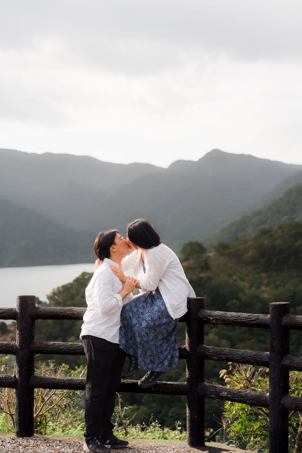 A romantic couple in white shirts share a tender moment by a wooden fence overlooking misty mountains and a lake.