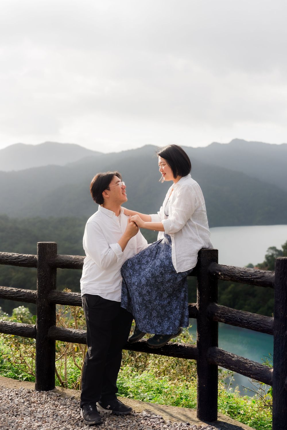 A couple in white shirts share a tender moment by a wooden fence overlooking misty mountains and a lake.