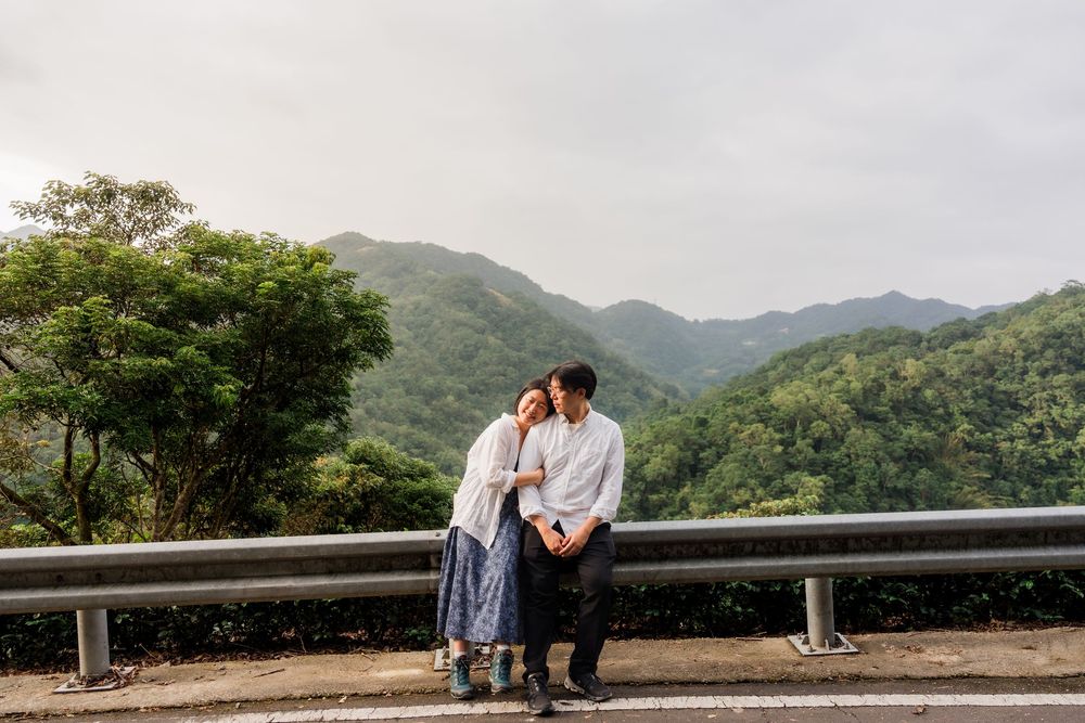 Couple embraces while standing on mountain overlook with scenic forest valley views in the background.