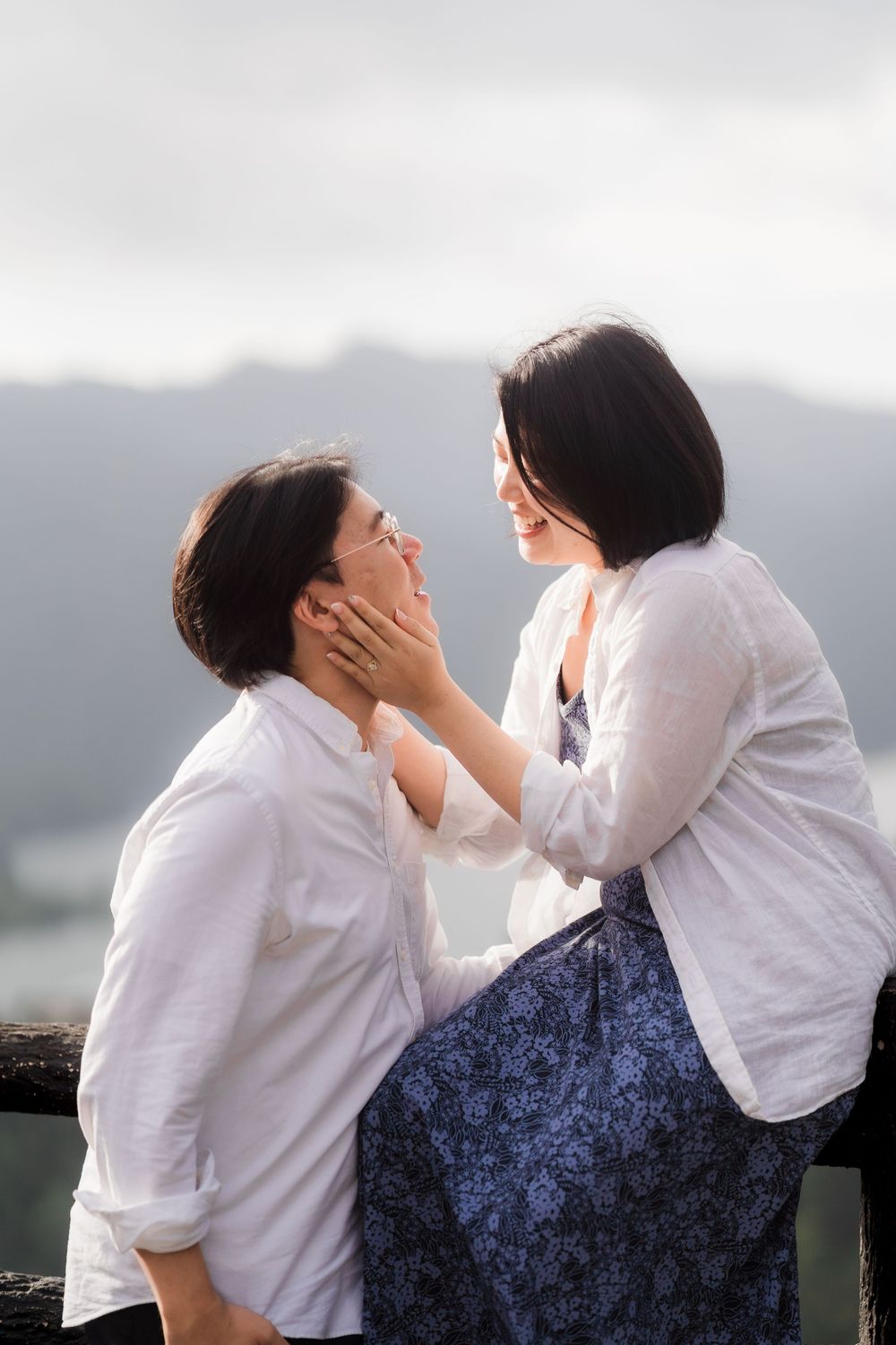 Two people in white shirts share an intimate moment against a scenic mountain backdrop with wooden railings.