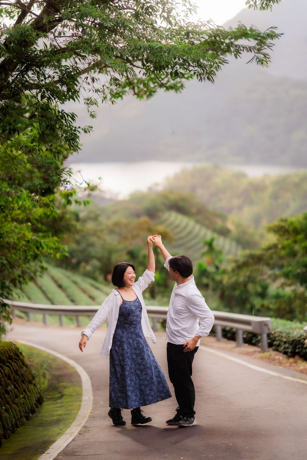 A couple dancing together on a winding mountain road surrounded by lush tea plantations and misty scenery.