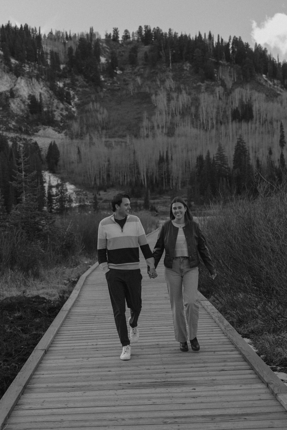 A playful couple runs and laughs together on a wooden boardwalk path through mountain wilderness.