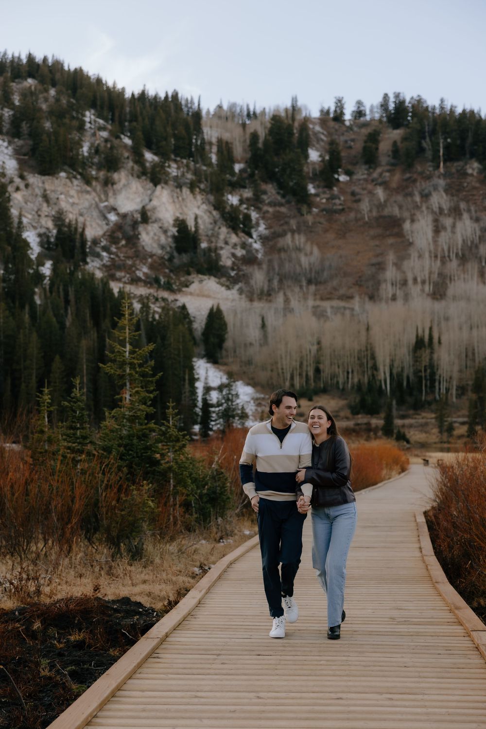 A couple walks hand in hand along a wooden boardwalk path through a mountainous landscape with pine trees.