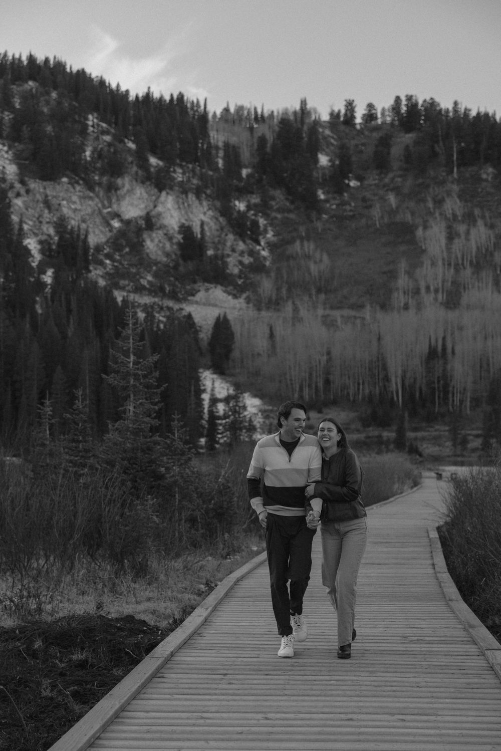A couple walks along a wooden boardwalk path through a scenic forest landscape with evergreen trees in the background.