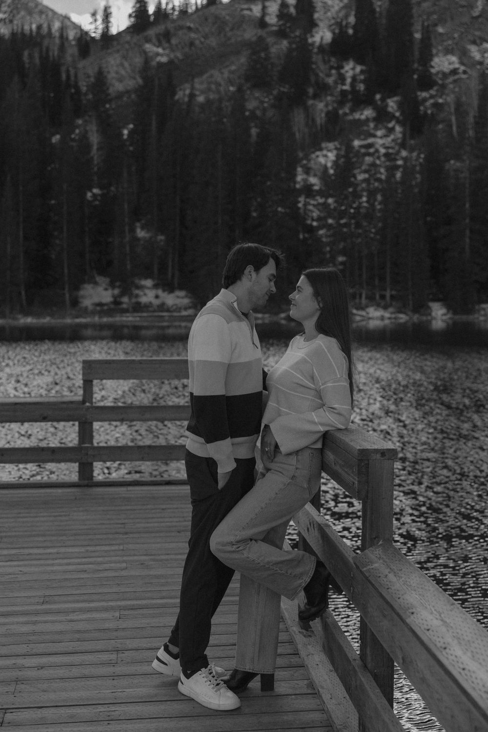 A couple shares a romantic moment on a wooden deck with snow-capped mountains and evergreen trees in the background.