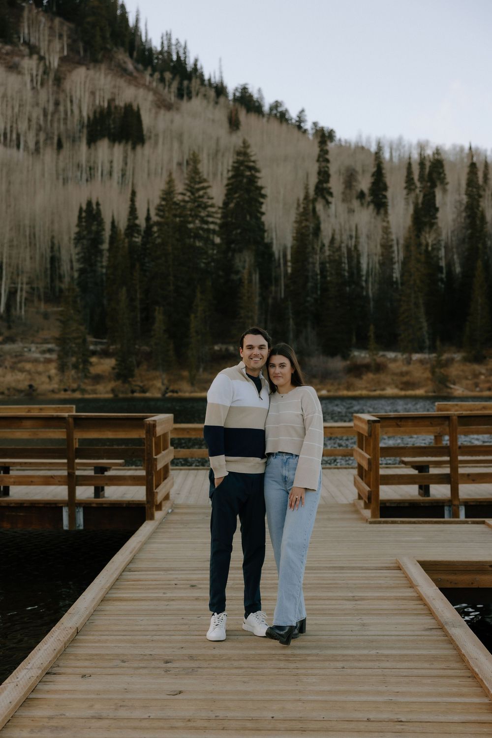 A wooden boardwalk extends over a lake surrounded by evergreen trees and mountains in a serene natural setting.