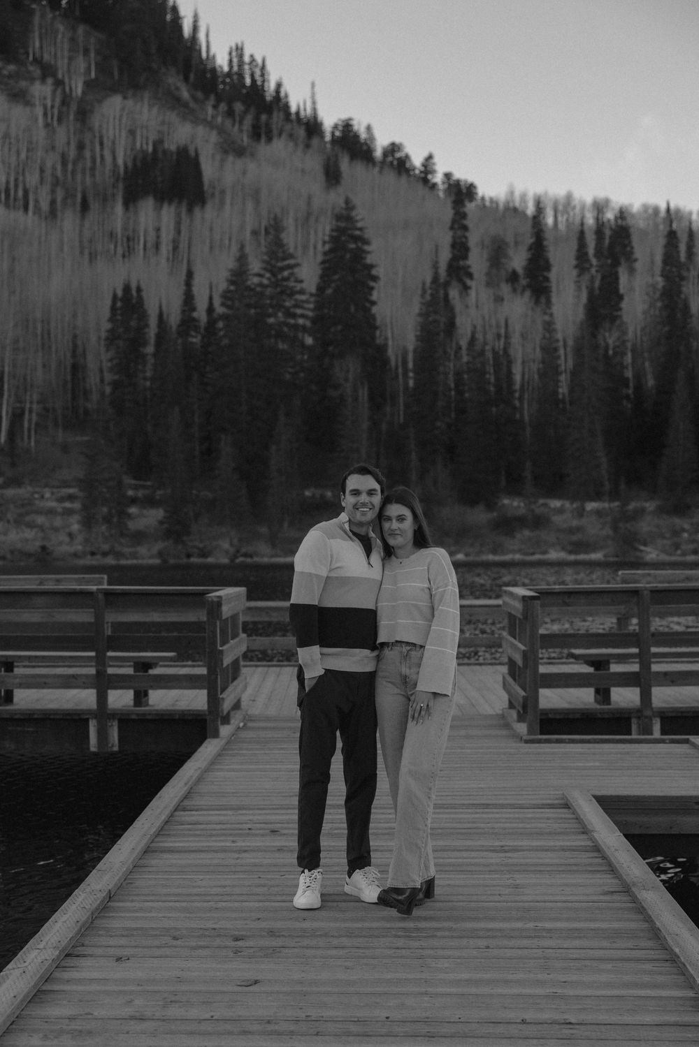 A couple stands together on a wooden dock with evergreen forest and mountain slopes in the background.
