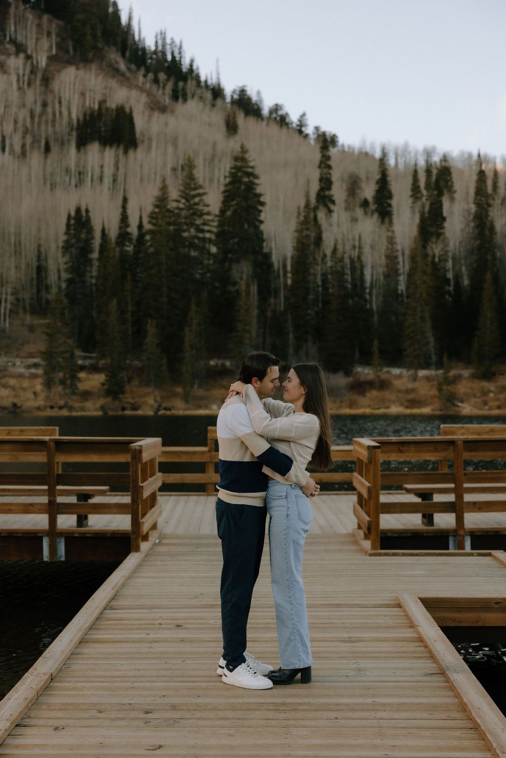 A couple shares an intimate moment on a wooden deck overlooking a scenic mountain lake surrounded by evergreen trees.