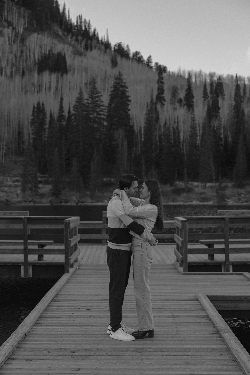 A romantic couple shares an intimate dance moment on a wooden dock surrounded by evergreen trees in black and white.