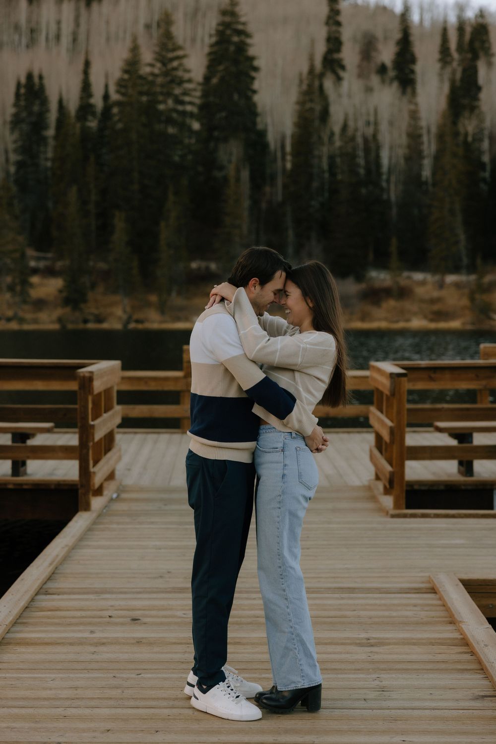 A couple embraces tenderly on a wooden boardwalk surrounded by evergreen forest and mountains in the background.