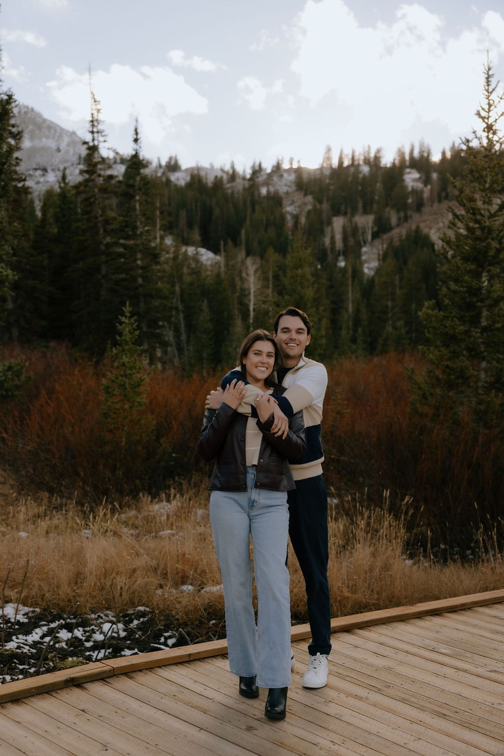 Two people share a tender moment together on a wooden deck surrounded by evergreen trees and mountain scenery.