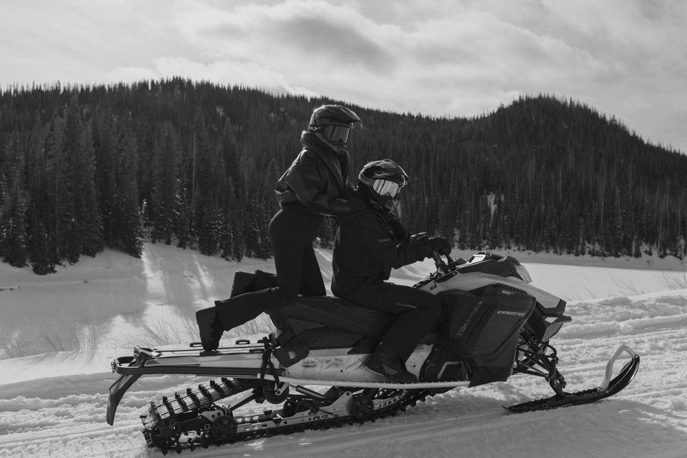 Snowmobiles speeding through deep powder snow with evergreen forest and mountains visible in the distance.