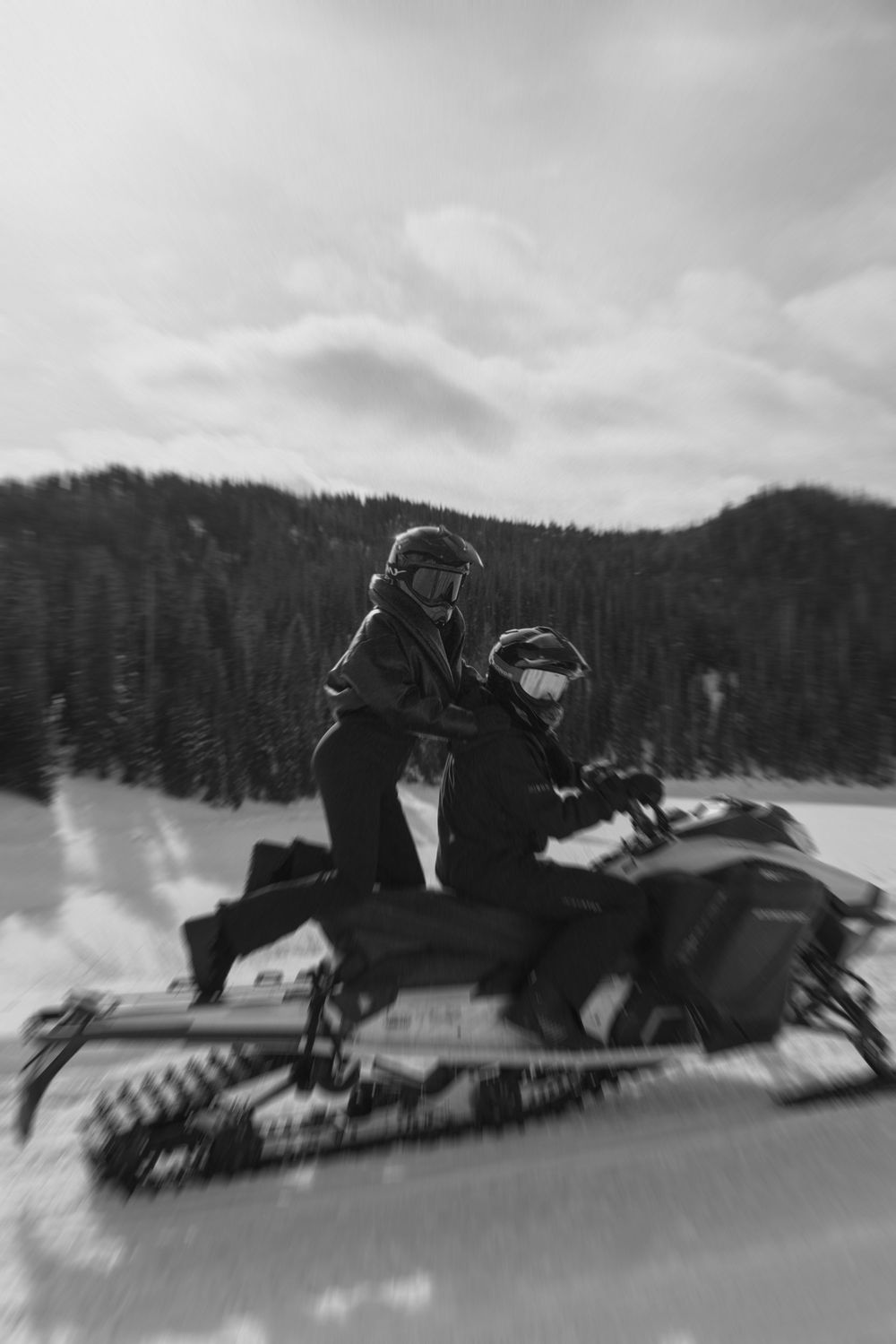 A sequence of snowmobile riders racing across a snowy landscape with evergreen trees in the background.