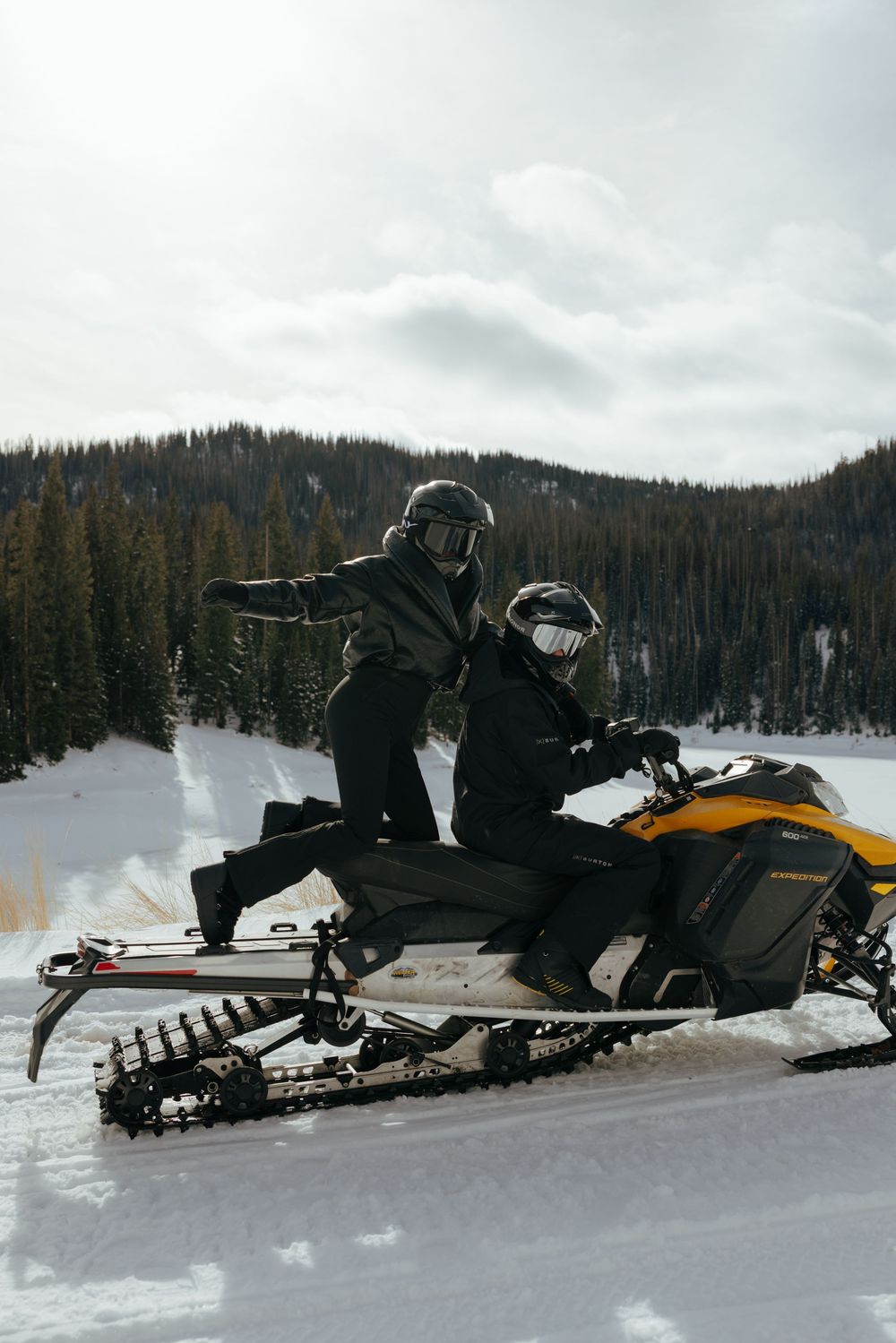 Two snowmobilers pose on their yellow snowmobile against a scenic winter mountain backdrop with evergreen trees.
