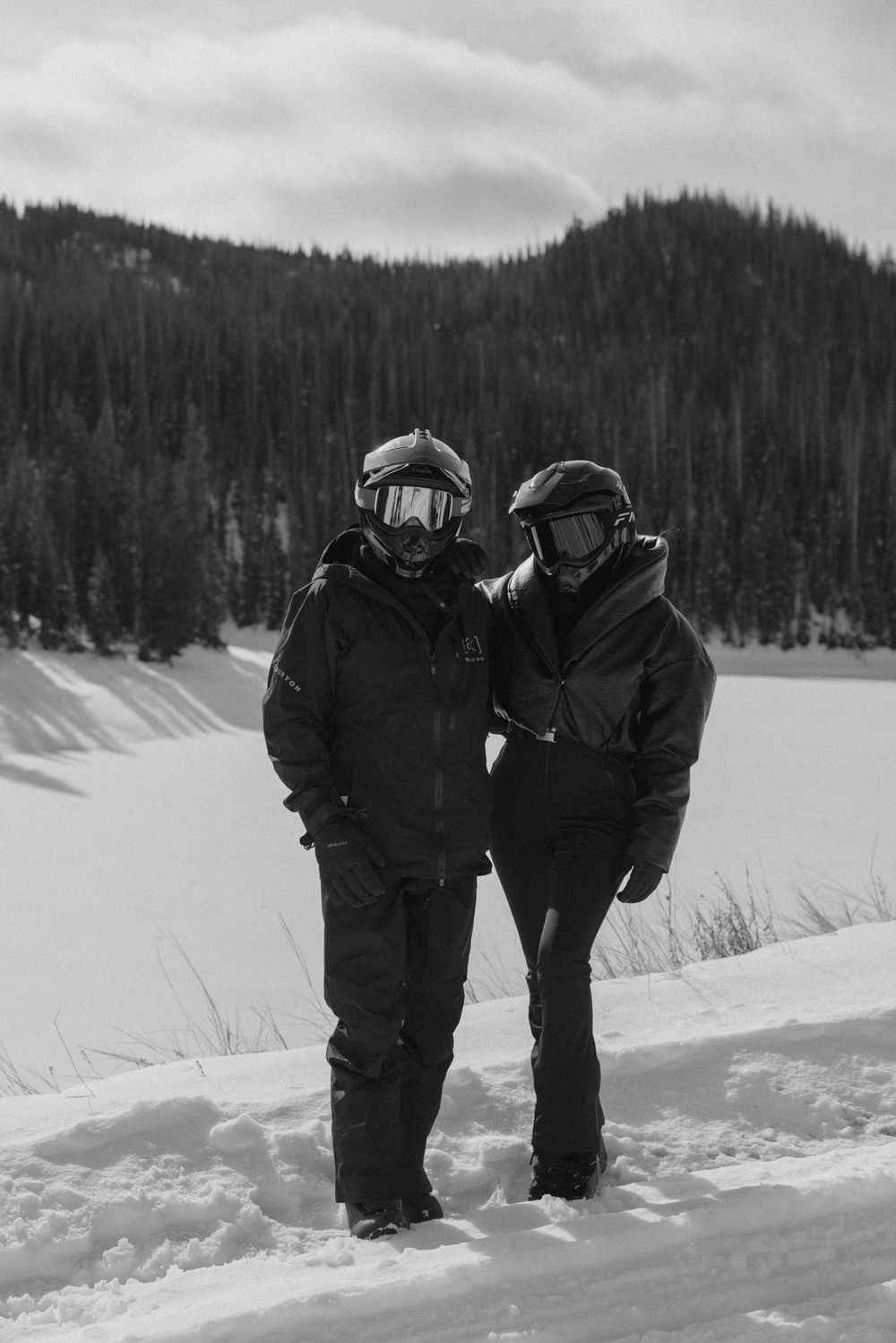 Two winter sports enthusiasts stand together in deep snow with a snowy mountain landscape and forest behind them.