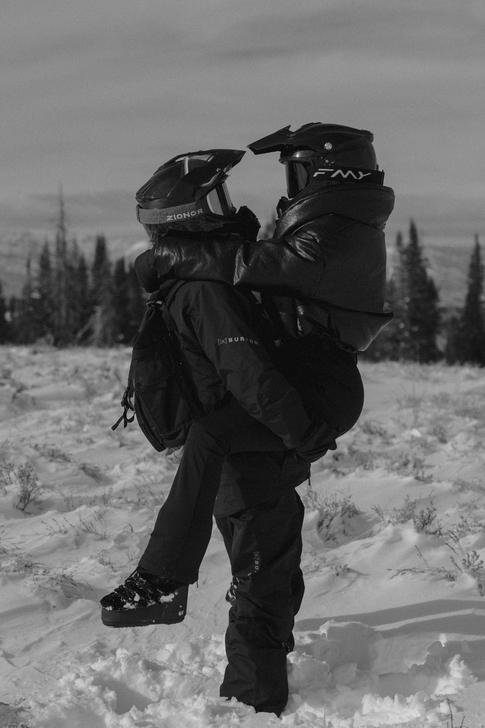 A person in winter gear bends down to examine snow conditions in a snowy forest landscape.
