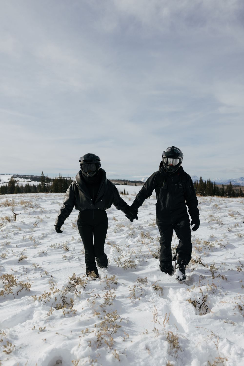 Two people in dark winter clothing walk through a snowy field on an overcast day.